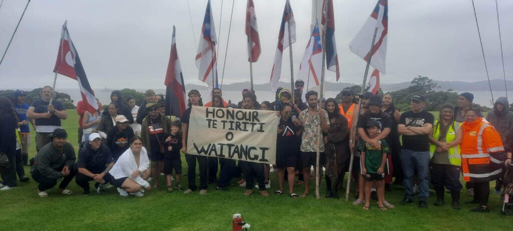photo of protest that went from the Waitangi lower grounds to the upper grounds. People are holding flags and have a banner that reads "HONOUR TE TIRITI O WAITANGI"