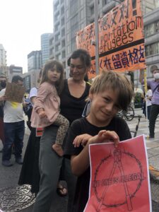 family holding signs that read "no one is illegal" japanese signs against deporting refugees in the background.